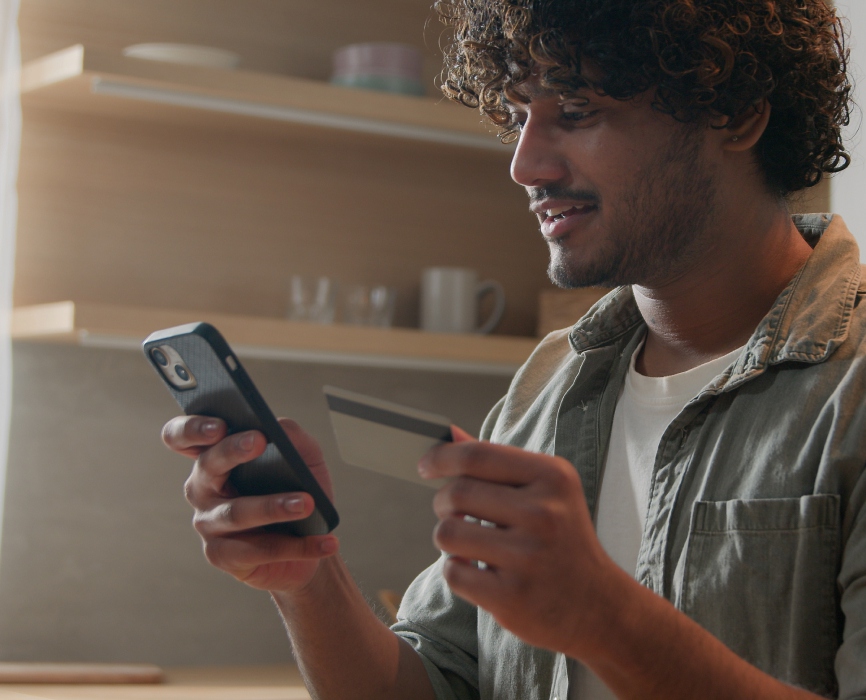 Happy man with phone and credit card using instant mobile payment at home kitchen Indian guy male customer shopper buyer buying online purchase smartphone shopping food delivery e-banking app