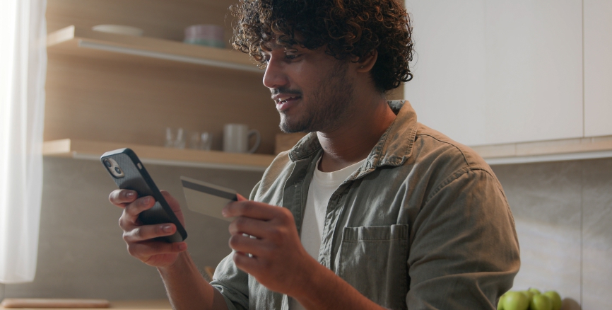 Happy man with phone and credit card using instant mobile payment at home kitchen Indian guy male customer shopper buyer buying online purchase smartphone shopping food delivery e-banking app